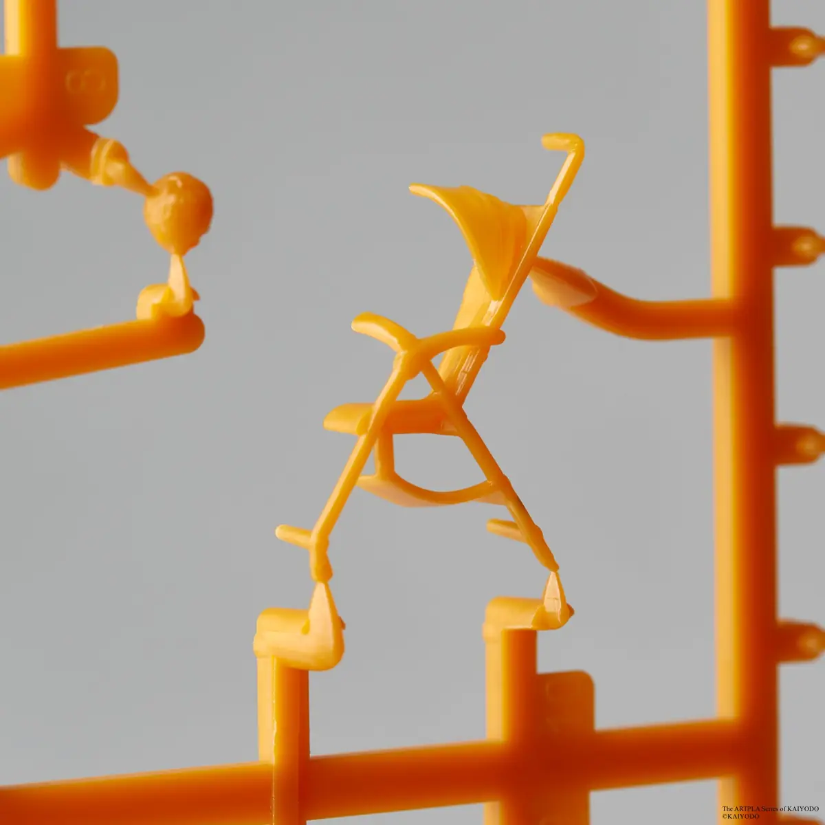 Orange miniature folding-chair component attached to a plastic runner against a gray background