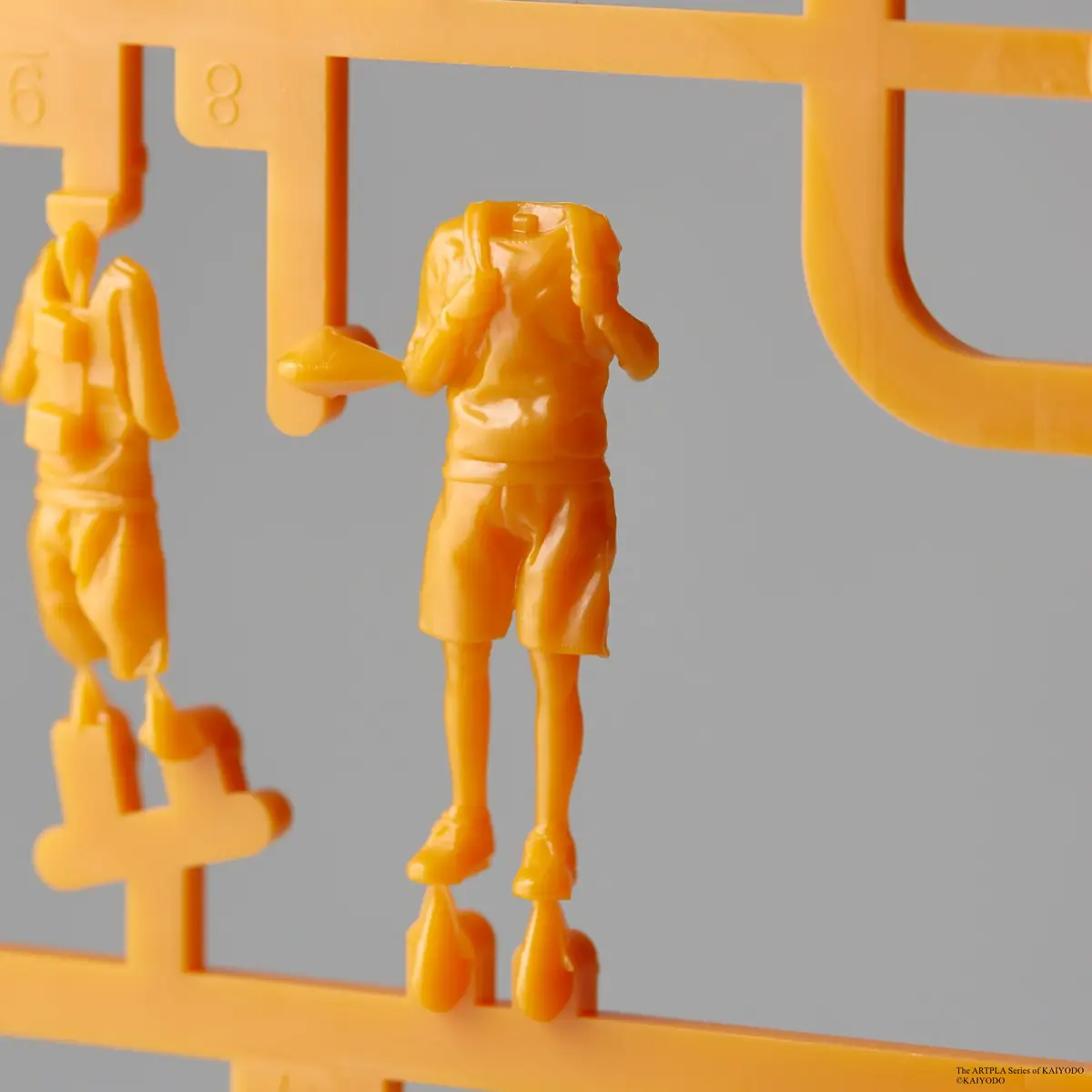 Two orange miniature human body components attached to a plastic runner against a gray background